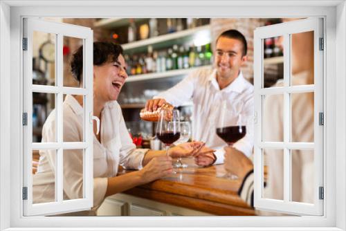 Two smiling female friends sitting in the bar and drinking red wine while bartender pouring some drink